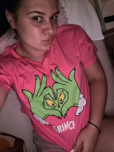 In a dimly lit room, a young woman with dark hair pulled back and wearing a bright pink t-shirt with a Grinch graphic lies on a bed. The t-shirt features two green, clawed hands of the Grinch forming a heart shape, with the word "GRINCH" printed below. The woman is looking directly at the camera with her lips pursed. She is wearing gray shorts. The background shows a white pillow and a dark, indistinct area which could be furniture or a wall. There are no discernible elements that can be used to determine the location within the city of Rogosh, Bulgaria.