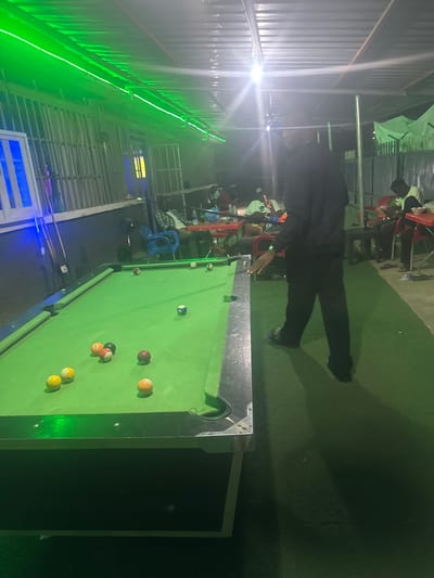 The image depicts an outdoor pool hall in Karu, Nigeria. The pool table is green with multiple pool balls positioned on it. A person dressed in black stands at the edge of the pool table, presumably preparing to play. In the background, there are people sitting on chairs, some near tables, possibly watching the game or socializing. The area is illuminated by artificial lighting, including green lights along the roof or structure. The environment appears to be a casual social gathering spot.