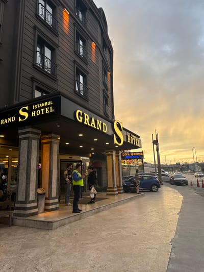 The scene depicts the street-level entrance of the Grand S Istanbul Hotel in Esenler, Turkey, under an overcast sky with a prominent golden-orange sunset hue.

The five-story hotel building, constructed of dark grey material, features rectangular windows and black wrought-iron balconies on its upper floors. Orange light from the sunset reflects distinctly on the undersides of some upper-floor window sills. The hotel's entrance is marked by a prominent black canopy with illuminated yellow lettering displaying "GRAND S İSTANBUL HOTEL." Beneath this, a smaller illuminated sign advertises "DİVA İŞKEMBE RESTAURANT." The entrance is supported by four square pillars with light and dark horizontally striped marble-like cladding. A revolving glass door is visible behind the pillars.

On the raised entrance platform, three male individuals are present. One man in a blue top and a bright yellow reflective vest stands facing right, appearing to converse with a man to his immediate right (viewer's left) who is wearing a plaid jacket and jeans. Another man, to the immediate left of the man in the vest (viewer's right), wears a dark top and holds a white plastic bag. A grey metal trash receptacle is positioned near one of the pillars.

Adjacent to the hotel, a multi-lane asphalt road runs parallel. A blue hatchback vehicle is parked close to the hotel's lower pavement level. A female individual in a patterned dress stands by the rear of this blue car, facing away from the camera. Several other vehicles, including a dark sedan, are present on the road, with some in motion. Further down the road, two vertical flagpoles stand, and orange traffic cones are visible along the roadside. In the distance, a concrete overpass or bridge structure is visible, consistent with a highway environment. The ground immediately in front of the hotel consists of light-colored tiled pavement, transitioning to a raised platform at the entrance.