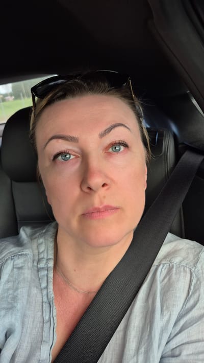 A woman with light skin and blue eyes is sitting in the driver's seat of a car, facing the camera. She has blonde hair pulled back and is wearing sunglasses on top of her head. Her left eye appears red and slightly swollen. She is wearing a light blue collared shirt and a silver necklace. A black seatbelt is visible across her chest. The background shows the interior of a car and a blurred outdoor scene with greenery and a building in the distance. The image was taken in Hærland, Norway.