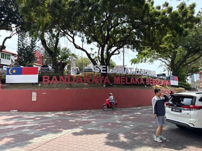 The image captures the "Selamat Datang Bandaraya Melaka Bersejarah" sign, welcoming visitors to the historic city of Melaka, Malaysia. The sign is adorned with Malaysian flags on either side and is set against a red brick wall with large trees looming above. A man stands beside a white car with license plate "ANB 8832," while a motorcyclist drives past in the background. The scene is captured during the daytime under bright sunlight.