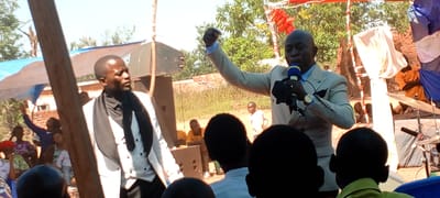 The image depicts a gathering, possibly a church service or event, in an outdoor setting. It is daytime with bright sunlight. In the foreground, individuals are looking at the camera.
A man in a white suit holds a microphone. Beside him stands another man in a white vest and dark clothing. Both appear to be the focal points of attention.
In the background, a crowd is seated. The setting suggests a makeshift outdoor venue, with tent-like structures erected using wooden poles and draped fabric. Musical instruments are visible on the right, indicating live music.
The location is Kalemie, Democratic Republic of the Congo, based on the visual characteristics of the scene.