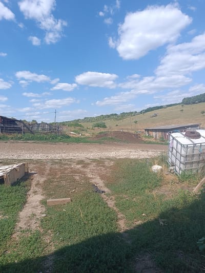 This is an outdoor, eye-level, full-shot photograph of a rural landscape. The scene appears to be a farm or homestead in Luminița, Moldova. The sky is a bright blue with scattered white cumulus clouds. The foreground is dominated by a dirt path with patches of dry grass and some scattered debris, including a light-colored wooden structure. To the right, a large white plastic tank with a metal cage is visible, alongside a smaller white bucket. Behind the tank are some weathered wooden structures, possibly sheds or outbuildings, with corrugated metal roofs. A wooden fence runs across the middle ground, separating the homestead area from a grassy, undulating hillside. The hillside is covered in dry grass with scattered trees and shrubs. In the far distance, a line of trees marks the horizon. The overall impression is one of a rustic, somewhat unkempt rural setting on a sunny day. There are no people or animals visible in the frame.