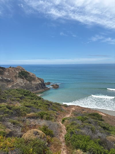 This is a high-angle, landscape, vertical shot of the Odeceixe coastline in Portugal. The image captures a dramatic view of the Atlantic Ocean meeting a rugged, vegetated cliff face. The water is a deep blue with white waves crashing near the shore, and a sandy beach is visible to the right. The cliff is covered in dense, green and brown scrub vegetation, with a dirt path winding its way along the edge. In the distance, the ocean stretches out to a hazy horizon, with a few scattered rocks emerging from the water near the cliff. The sky is a clear, bright blue with wispy white clouds on the left side. The lighting suggests it is a sunny day, likely midday or early afternoon. There is no visible text or people in the image, and the overall mood is one of natural beauty and serene isolation.