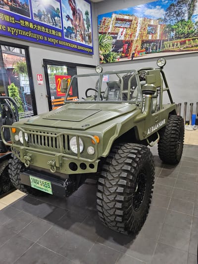 An olive green, open-top, militaristic-style vehicle is parked on a dark gray tiled floor inside what appears to be a showroom or museum. The vehicle has large, aggressive-tread tires, exposed suspension components, a roll cage, and a snorkel exhaust. The front of the vehicle features a reinforced bumper with tow hooks, round headlights, and a slatted grille. A green license plate with white Thai characters and the number "5555" is visible on the front. Text on the side of the vehicle reads "AI BAAN HAWK". In the background, several large framed artworks are displayed on a gray wall. One artwork depicts dinosaurs, and another shows a colorful building decorated with many lanterns and ornaments. The environment suggests an indoor exhibition space, possibly related to themed attractions or car shows in Na Chom Thian, Thailand.