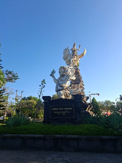 This is a low-angle, eye-level, outdoor, full shot of a large, white stone statue located in the city of Gianyar, Indonesia, under a clear, bright blue sky. The statue depicts a mythological scene with a large, imposing figure at the bottom, possibly a demon or rakshasa, holding an object in its right hand. Above this figure, a smaller, more graceful figure, likely a deity or a prince, is seated on its shoulders, with large, spread wings. The statue is mounted on a dark stone pedestal with a plaque that reads "TAMAN KOTA GIANYAR" in English and a script below that appears to be Balinese. The statue is surrounded by lush green manicured bushes and plants, with some taller plants and trees in the background. To the left, there are some more trees and a utility pole with wires. In the distance, a construction crane is visible. The lighting suggests it is either early morning or late afternoon, with the sun casting a warm glow. The scene appears to be a public park or square, a landmark within the city of Gianyar.