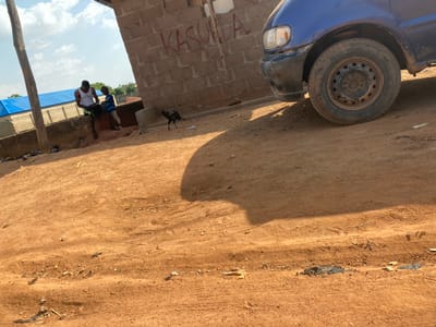 The image, set in Hayin Banki, Nigeria, displays an outdoor scene featuring a dirt road, structures, people, and a vehicle under a bright, clear sky.
A wide, unpaved dirt road dominates the upper and left sections of the frame, showing numerous tire tracks and scattered refuse, including plastic items. A large, dark shadow stretches across a portion of this road.
In the lower left, the front part of a blue car is visible, featuring a single headlight and a dirty, dark grey steel wheel. The vehicle appears to be stationary. Adjacent to the car, and occupying the lower right side, is a concrete block wall. Red graffiti is scrawled on this wall, with the word "KASUWA" clearly discernible. Above the wall, a small, dark-feathered bird, possibly a chicken, is perched on a ledge.
Two adult males are present in the mid-ground on the right. One, wearing a white sleeveless shirt, stands near the wall. The other, partially visible, wears a blue and white striped shirt. They appear to be resting or observing. Behind them, a vibrant blue tarp is visible, possibly forming a temporary roof or market stall, supported by wooden poles. The background reveals a light blue sky with scattered white clouds. The strong, angled shadows suggest either mid-morning or late afternoon. The scene captures an ordinary moment in a semi-urban or rural environment.