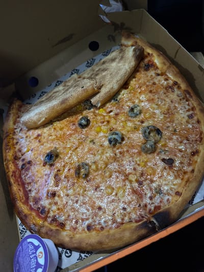 A partially eaten pizza sits in a cardboard box. A slice is detached and lies alongside the remaining pizza. The pizza is topped with cheese, corn, and what appear to be sliced black olives. A small container of garlic and herb dip is visible in the lower right corner of the box. The box indicates "Design Fire Eat" and "Takeaway" on its sides. The exact location in Rushcliffe, United Kingdom, cannot be determined from the image. No people are visible.