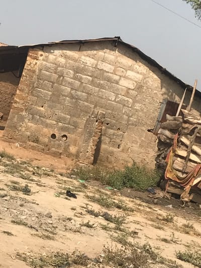 The image displays a scene in Ago-Abo, Nigeria, dominated by a single-story building constructed from unrendered concrete blocks. The visible wall exhibits significant weathering, discoloration, and minor structural wear, including a vertical section of exposed reddish brickwork and two circular holes near its base. The roof is composed of rusted, uneven corrugated metal sheets. To the right of the building, a large stack of cut timber or wooden planks is present, partially obscured by reddish-brown fabric or tarp, with a rudimentary wooden ladder leaning against it. The ground in the foreground and mid-ground is dry, light-brown earth with sparse patches of low green vegetation. On the ground, directly in front of the building, a pair of light-green flip-flops and a dark, shapeless object are visible. A light-colored chicken is partially visible on the far right, near the stacked timber. The sky overhead is a clear, light blue. No human subjects are present in the scene.