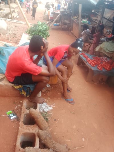 The image captures a vibrant street market scene, likely in a rural or semi-urban setting, characterized by red-brown dirt ground and rustic market stalls. The lighting suggests it is daytime, with natural light illuminating the area.
In the foreground, two young people are resting. On the left, a person wearing a red t-shirt and black shorts is seated barefoot on a low structure made of concrete blocks. They are facing away from the viewer, towards the right, with their hands clasped over their head, giving an impression of contemplation, weariness, or distress. A patterned cloth is placed on the concrete block beneath them. Nearby on the ground are two large, elongated brown root vegetables, resembling yams, along with a small green and white packet (possibly cigarettes or a similar product) and a bundle of white string or wire. A light blue tarp is visible behind this person.
To the right, another person, also wearing a red t-shirt over blue shorts, is seated, possibly on a small yellow container. This individual is looking down and smiling or laughing, with one hand on their lower leg. They are wearing blue flip-flops. A black headband with the white text "NIKE" is worn on their head.
In the midground and background, the market activity unfolds. To the immediate right of the second person, there's a wooden market stall with a bountiful display of bright red tomatoes laid out on a blue fabric. Further back, more market stalls are visible, constructed from wood and corrugated metal, and various items are displayed for sale. A large pile of green leafy vegetables sits on the ground between the two foreground subjects and the background stalls. Several other people, including adults and children, can be seen moving around the market, some walking, others near the stalls, contributing to the bustling atmosphere. A bamboo pole is noticeable on the left side, extending upwards.