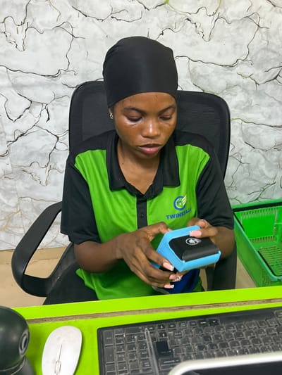 A woman in a green and black uniform with the "TWINSFAJA" logo is sitting at a desk holding a blue handheld device. The desk has a dirty keyboard, a mouse, and other items on it. There is a green basket in the background. The location is Ijegun, Nigeria.