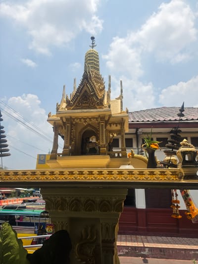The image shows a spirit house, a small shrine, likely located in Phra Nakhon Si Ayutthaya City Municipality, Thailand. The spirit house is ornate, constructed of light-colored stone or plaster, and decorated with gold leaf details. It has a multi-tiered roof culminating in a spire. Inside the shrine, a small figurine is partially visible. A black vase sits on top of the shrine with plant offerings. A garland of orange flowers hangs on the side. In the background, the building has a tiled roof and red walls. The sky is blue with scattered clouds. Power lines run across the frame. Parts of the street, including people, are visible in the background, however these parts are out of focus.