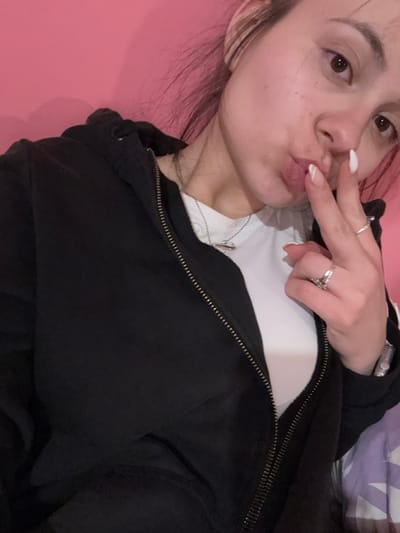 The image displays a female individual situated indoors, identified as being in Radomir, Bulgaria. She has light skin, brown eyes, and light brown hair. The subject is wearing a black hooded jacket with a brass-colored zipper, partially unzipped, over a white t-shirt. A silver-colored necklace with a small pendant is visible. Her left hand is raised, with the index and middle fingers extended and touching her pursed lips, creating a "V" shape. Her fingernails are long, almond-shaped, and painted with white French tips. Two silver-colored rings are present on her left middle and ring fingers, and a watch or similar accessory is visible on her left wrist. The background consists primarily of a solid pink wall, with a blurry purple and white patterned object visible in the lower right portion of the frame.