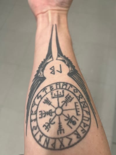 The image shows a close-up of a tattoo on a person's forearm. The tattoo features a compass-like design with Norse runes and what appear to be stylized wings extending upwards, forming a bird-like silhouette. The background is a plain, neutral surface, suggesting an indoor setting. There are no identifiable landmarks or features that would indicate the location as Thessaloniki Municipal Unit, Greece.