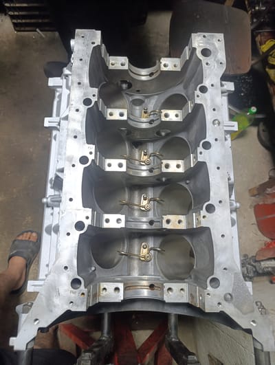 An overhead, eye-level, medium shot captures a four-cylinder engine block, likely an aluminum alloy, positioned on a red metal engine stand. The block is silver and shows machined surfaces. The lower portion of the engine block is in focus, revealing the cylinder bores and the main bearing caps secured by bolts. Some of the bearing caps have clips attached to them. There are also various holes and mounting points visible on the exterior of the block. To the left, a portion of a bare foot with a dark sandal is visible at the bottom edge of the frame, indicating the presence of at least one person. The background is indistinct but appears to be an indoor workshop or garage environment with concrete flooring and some out-of-focus tools or equipment. The specific location in Novopavlovka, Kyrgyzstan cannot be determined from the image.