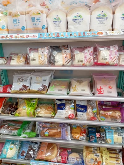 The image shows shelves stocked with various types of packaged bread in a store. The topmost shelf has white bread with blue and orange labels. Below that are packages of sliced bread. The third shelf contains bread with chocolate and strawberry flavors. The bottom shelves are filled with buns and other baked goods in clear plastic packaging. The packaging has a red label that says "10" suggesting it is a discount. There is a package with the number "65" on it. Based on the text on the packaging, the store is likely located in Kaohsiung, Taiwan.