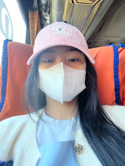 A young woman with long, dark hair wears a pink baseball cap and a white face mask, obscuring the lower half of her face. She is seated on a bus, indicated by the orange and blue patterned seat in the background, and the interior of the bus is visible above her. The woman is wearing a white knitted cardigan over a light blue shirt. A gold floral brooch is pinned to the left side of her cardigan. The overall scene suggests a casual outing or travel. The location in Sumber Suko, Indonesia, is not directly discernible from the visual information within the image itself.