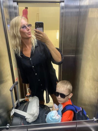 In the elevator of a residential building in Giachrypsh, Abkhazia, a blonde woman in a black open-collared shirt and sunglasses takes a selfie with her phone. Beside her, a young boy wearing sunglasses and an orange and blue rash guard with a blue backpack looks away from the camera. Luggage, including a black suitcase and a white bag, is visible in the elevator. In the reflection of the elevator doors, another person in a pink top is seen in the hallway.