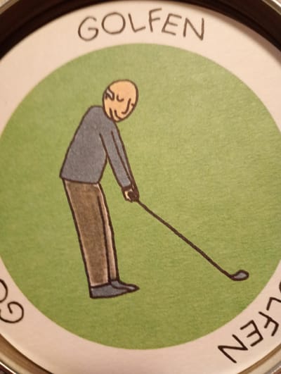 The image shows a close-up of a circular design on what appears to be the lid of a tin. The design features a cartoonish illustration of a person playing golf. The golfer is depicted with a bald head, a grey shirt, and brown pants, holding a golf club. The background is a solid green, representing a golf course. Above the illustration, the word "GOLFEN" is written in black capital letters. The word "GOLFEN" is also partially visible in a curved fashion along the bottom edge of the circular design. The image is not indicative of any specific location in the city of Rosenheim, Germany, as it is a generalized illustration.