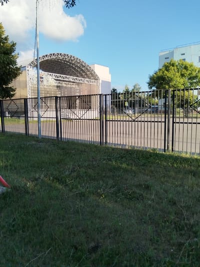 This media file captures an outdoor scene on a clear, sunny day, likely in the afternoon, given the shadows. The foreground is dominated by a grassy area and a black metal fence with vertical bars, interspersed with some decorative diamond shapes. Beyond the fence, a paved area is visible, leading to a stage structure with a curved metal roof supported by a lattice of trusses. The stage is enclosed by a semi-transparent material.

Behind the stage, a light-colored building with windows is visible, and further in the background, a taller, pale blue building with multiple windows and a flat roof suggests an urban or suburban environment. Lush green trees are scattered throughout the scene, with one particularly prominent tree to the right of the stage structure. A lamppost adorned with decorative lights stands to the left of the stage.

No people are clearly visible, and there is no discernible activity taking place. The overall impression is one of an outdoor public space, possibly a park or a recreational area, with a dedicated stage for performances or events. The environment appears well-maintained and serene. There is no visible text in the image. The location context provided is Рощинский, Russia.
