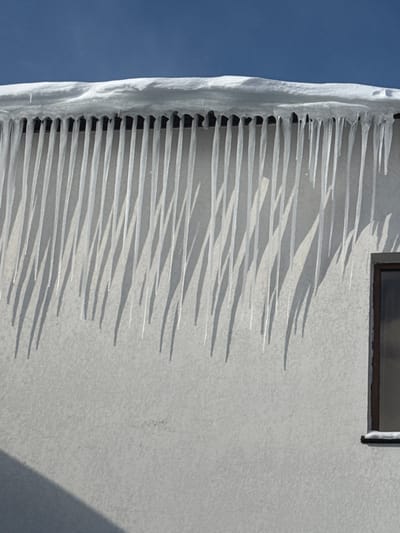A building's corner shows icicles hanging from the roof's edge, casting distinct shadows on the white exterior wall. A dark-framed window is visible near the top corner of the structure. The icy fringe has varying sizes and shapes. The sky is blue in the background, accentuating the stark white of the building's facade.