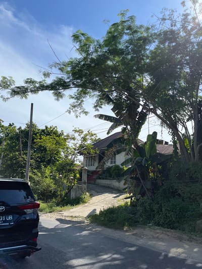 The image depicts a street scene in Kota Kuala Simpang, Indonesia, during daytime. The perspective is from within a vehicle, looking forward and slightly upwards. Lush green trees and foliage dominate the left side of the frame, with branches hanging over the road and obscuring parts of the sky. A house with a white exterior and a dark, sloped roof is visible nestled among the trees. Power lines crisscross the scene, suggesting an inhabited area. The road itself appears to be paved and runs along the right side of the image, with a dirt and gravel shoulder. Shadows from the trees are cast across the road, indicating it is likely midday or afternoon.

In the foreground, the rear of a black car is visible, with its taillights and license plate in view. The license plate displays the text "31 UB" and "127". The overall impression is one of a tropical, residential or semi-rural setting on a sunny day. The bright sunlight filtering through the leaves and the clear blue sky suggest fair weather. The scene appears calm and ordinary, with no specific activity or event being the primary focus beyond the everyday existence of a street in this location.