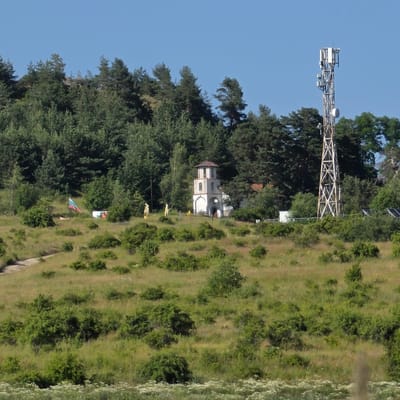 This image shows a landscape with a telecommunications tower and a small church. The scene is set in Ravnogor, Bulgaria. The foreground features a grassy, hilly terrain with scattered bushes and some wildflowers. A dirt path is visible on the left side, winding its way up the hill. In the middle ground, on a slight elevation, stands a white church with a red roof, its architecture suggesting it might be a belfry or a chapel. To the left of the church, a Bulgarian flag is displayed on a flagpole, with a few scattered items and figures that appear to be human statues or mannequins. Further to the right, a tall, lattice telecommunications tower is prominently positioned against a clear blue sky. The background is dominated by a dense forest of evergreen trees. The overall impression is of a rural, possibly mountainous, area.