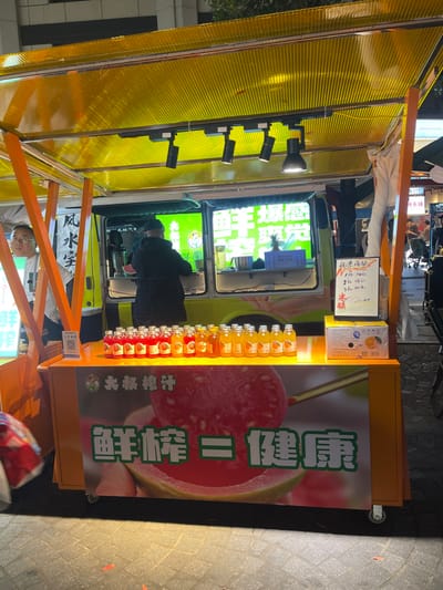 A yellow food cart with a yellow awning sits on a sidewalk in Hang Zhou Shi, China (latitude: 30.264, longitude: 120.156). The cart is displaying rows of small glass bottles containing a bright red liquid, possibly guava juice. The bottles are arranged in front of a large, colorful advertisement featuring a guava fruit with the Chinese characters "Guava = Health" on a yellow background. A man wearing a black jacket is standing beside the cart. The cart is parked in front of a larger, white food truck with a yellow sign that reads "Guava" and depicts a picture of a guava fruit. The truck has a window and interior that is partially visible and appears to be stocked with various items. The scene is illuminated by streetlights, casting a yellow glow on the sidewalk.
