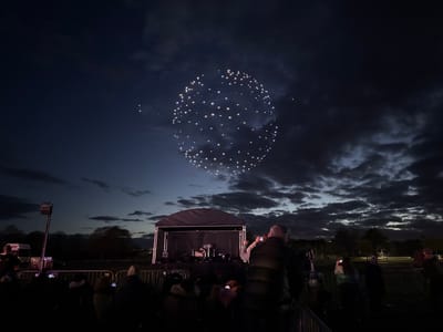 The image depicts a dark outdoor event, likely a concert or performance, in what is possibly South Norfolk, United Kingdom. A stage with a black canopy is visible in the middle ground, containing a drum set and other equipment. A crowd of people stands in the foreground, with many holding up phones to record the event. Above the stage, in the night sky, is a formation of lights arranged in an oval or circular shape. The sky is filled with dark, textured clouds. The overall lighting is dim, creating a nighttime atmosphere. The presence of a truck and other equipment suggests that this event is taking place in a park or open space. A metal barrier separates the crowd from the stage.