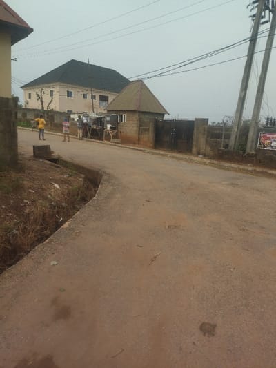The image shows a street in New Karu, Nigeria, located at latitude 8.97 and longitude 7.551. The street is paved with asphalt, and there is a small drainage ditch on the left side of the street. The street is empty, but there are a few people in the distance. They are standing in front of a building on the right side of the street. The building appears to be a residential home with a gate. There are power lines running above the street. The sky is overcast, and there are a few clouds in the sky. There are trees and vegetation along the sides of the street.