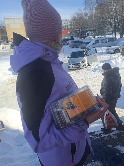 The image displays a clear winter day scene in Bolshaya Elhovka, Russia. In the foreground, a person is viewed from behind and slightly to the right, wearing a light purple knit beanie and a purple and black winter jacket with a visible fur collar. This individual holds a clear plastic container, bearing a label with Cyrillic text including "ТОРТ" (CAKE), in their right arm; their fingernails are painted a light pink. The ground is heavily covered in snow, with some areas of visible asphalt. In the mid-ground, a white Chevrolet Aveo sedan is parked on the snow, its driver's side door open. The vehicle's Russian license plate contains the characters "K 471 CX 71". To the right of the Chevrolet, another person, seen from behind, walks rightward, wearing a dark winter jacket with a fur-lined hood and carrying a red and white plastic bag in their left hand. The background features several other parked vehicles, including a white van partially covered in snow, and multi-story buildings, one with a light yellow and white facade, under a clear blue sky. Bare deciduous trees are visible in the distance.