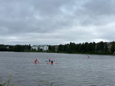 The image captures a wide shot of the city of Lysva, Russia, focusing on a lake with several people engaged in water activities. The sky is overcast with clouds. In the foreground, on the lake, there are three individuals paddleboarding. One person is further to the left, another is in the center, and a third is to the right. All are wearing orange life vests. The person in the center is paddling a blue board, while the other two are on red boards. There is a yellow buoy visible on the water between the leftmost paddleboarder and the far shore. The middle ground shows a line of trees bordering the lake, behind which are residential buildings, some with green roofs. The architecture suggests a residential area. The overall scene depicts a recreational day on the water in Lysva.