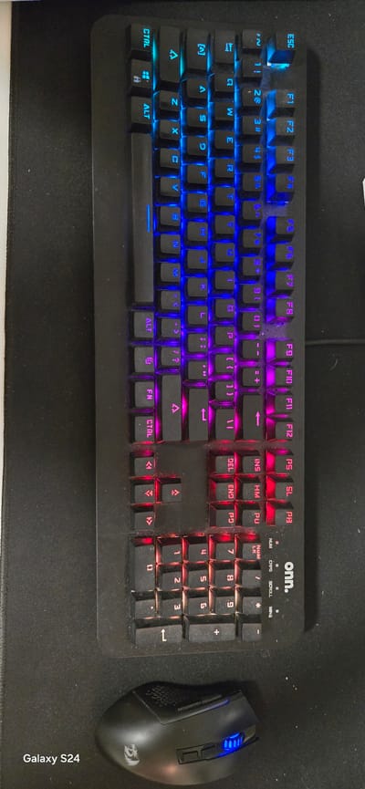 A top-down shot captures a black computer keyboard and a black gaming mouse resting on a dark fabric mouse pad. The keyboard, branded "onn.", features vibrant RGB backlighting, transitioning from blue on the left side (including the ESC and F1-F4 keys), to purple in the central section (F5-F12, alphanumeric keys), and red on the right-most sections (navigation keys, numeric keypad). Keys such as CTRL, ALT, Shift, Tab, Caps Lock, Enter, and Backspace are clearly visible, along with all alphanumeric keys, function keys (F1-F12), and media/control keys like INS, DEL, HOME, END, PG UP, PG DN, PRT SC, SCR LK, and PAUSE BRK. Indicator lights for NUM, CAPS, and SCROLL lock are present above the numeric keypad. Dust and small particles are visible on and around the keyboard keys, indicating use.
To the right of the keyboard, a black gaming mouse, bearing a faint Redragon logo on its side, displays blue LED lighting on its scroll wheel and DPI buttons. The mouse is positioned to the right of the keyboard, as if in active use. The mouse pad is dark gray or black. Along the bottom right edge of the mouse pad, the text "Galaxy S24" is visible vertically, suggesting the device used to take the photograph. The scene depicts an indoor, static setup with no direct activity taking place.