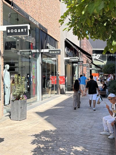 The image shows a pedestrian walkway in a commercial area in Ashdod, Israel, likely a shopping street or promenade. A building with brick and glass facades lines the left side of the walkway, featuring storefronts for various brands. Prominent signage includes "BOGART" displayed on the left storefront, with mannequins and merchandise visible through the glass. Further down the street, other businesses like "Original's" and "ALDO" are indicated by their respective signs.
Several people are walking along the paved walkway, which is illuminated by sunlight, casting shadows from overhead foliage and structures. On the right side, a man is seated on a bench, looking at his phone. The overall scene depicts a sunny day in an urban commercial setting.