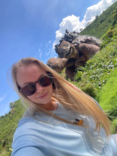 A blonde woman with sunglasses and long, blonde hair is taking a selfie outdoors. She is wearing a light blue t-shirt with a small yellow duck graphic on it. Behind her, a large, realistic sculpture of a tortoise is situated on a grassy hillside dotted with small white flowers. The background consists of a lush green, tree-covered slope under a bright blue sky with scattered white clouds. The scene appears to be a tourist attraction in Thôn 1, Vietnam.