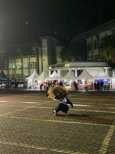 A man is performing a traditional dance in a paved parking lot in front of a large building in the city of Kecamatan Batu, Indonesia, at the coordinates -7.867 latitude and 112.514 longitude. The man wears a black shirt and pants, and a large, brown, fluffy headpiece. He is crouched down, with his arms extended, as if he is about to leap. The man's dance is performed in front of a building with white tent-like canopies set up in front of it. The canopies appear to be part of a market or festival. The building is two stories high and has many windows. The parking lot is empty except for the performer and a few people in the distance. The scene is illuminated by overhead lights. There are white lines painted on the parking lot. The building is in the background and the parking lot is in the foreground. The man is the only person in the foreground of the image. The night sky is visible in the background, and there are streetlights on the building.