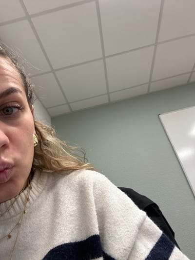 A selfie captures a woman indoors. A portion of her face is visible, featuring an eye with light-brown coloring and a small part of her nose and mouth. She wears a gold stud earring and layered necklaces. Her hair is curly and blonde. She's wearing a cream-colored sweater with thick, dark blue stripes. Behind her, there's a wall with square white tiles, a light-green wall, and a white rectangular board mounted on the wall. A black garment is partially visible behind her sweater.