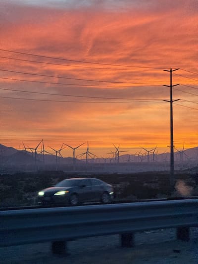 A landscape photograph taken in Desert Hot Springs, United States, shows a car moving on a highway with a field of windmills in the background under a vibrant orange and pink sunset. Overhead power lines stretch across the top of the frame, with a utility pole standing prominently on the right side. The guardrail of the highway is in the foreground, appearing dark and out of focus. The sky is a blend of orange and pink hues, with cloud formations visible. The silhouettes of numerous windmills are seen in the distance, suggesting a wind farm. A grey car with headlights on is passing by on the road. The overall scene conveys a sense of motion and energy, capturing the natural beauty of the sunset against the backdrop of a modern wind energy landscape.