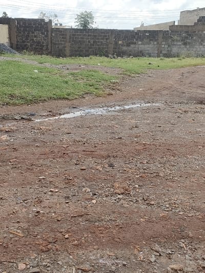 The scene depicts an outdoor undeveloped area in Juja ward, Kenya, under an overcast sky. The foreground and midground consist of a reddish-brown dirt and gravel surface, featuring a linear puddle of standing water, approximately 2-3 meters long, positioned horizontally across the mid-foreground. Scattered small stones and organic debris are present on the ground. A patch of green grass occupies the left midground. In the background, a low perimeter wall constructed of dark grey concrete blocks, reinforced with concrete pillars, extends horizontally. Behind this wall, on the left, a pile of grey construction aggregate is visible next to a light-colored structure. Further right, a small tree with light green leaves and several distant light-colored concrete buildings are observed, one featuring a white rooftop water tank. Overhead utility lines are faintly visible against the cloudy sky. No individuals or animals are present.