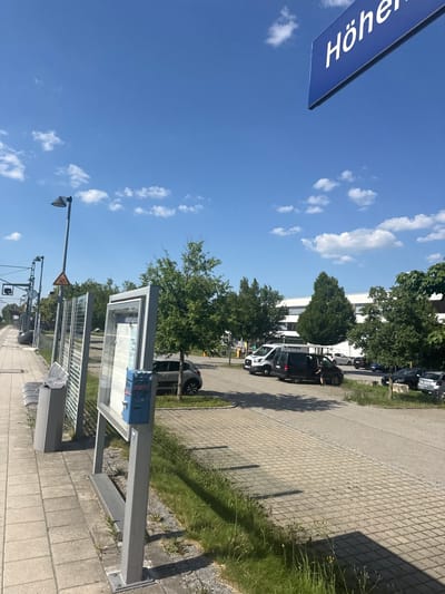 This is a photograph taken at a train station in Höhenkirchen-Siegertsbrunn, Germany. The scene is outdoors on a bright, sunny day with a clear blue sky dotted with fluffy white clouds. The perspective is from a low angle, looking upwards and diagonally across the scene.

In the foreground, a blue sign with white text is partially visible, reading "Höhen". To the right, a train station platform is visible, paved with interlocking concrete blocks. There are several modern station shelters with glass panels and metal supports. A waste bin is attached to one of the shelter structures. A street light with a distinctive lamp head stands on a tall pole. Further down the platform, there are more elements of the station infrastructure, including what appears to be a clock and traffic signals for trains.

Beyond the platform, there is a paved area with several parked cars. The cars appear to be a mix of passenger vehicles and a van, some of which have people inside or near them. A building with a white facade and dark window frames is visible behind the parked cars, bordered by lush green trees. The overall impression is of a quiet, functional public space on a pleasant day. There are no strong indications of specific events or emotions. The time of day is likely midday or early afternoon, given the bright sunlight and distinct shadows.