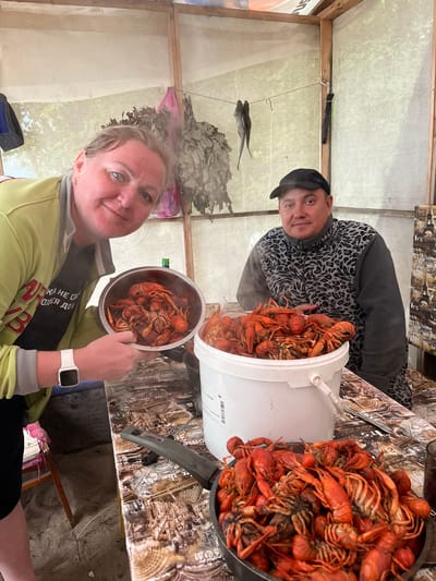 A woman and a man are standing behind a table covered with a patterned tablecloth. The woman, on the left, is holding a metal pot filled with cooked crawfish and smiling at the camera. She is wearing a light green jacket over a dark t-shirt with Russian text and a white smartwatch. The man, on the right, is wearing a black baseball cap and a grey fleece jacket with a leopard-like pattern. He is looking at the camera. In front of them, on the table, is a large white bucket overflowing with cooked crawfish, and a pan also filled with crawfish. Behind the individuals, there is a wooden structure with a sheer fabric covering, suggesting an outdoor or semi-outdoor setting, possibly in Derbeshkinskiy, Russia, as indicated by the presence of what appear to be birch whisks, commonly used in Russian saunas (banyas). There are also dried fish hanging from a line in the background. The overall atmosphere appears informal and celebratory, likely centered around the prepared food.
