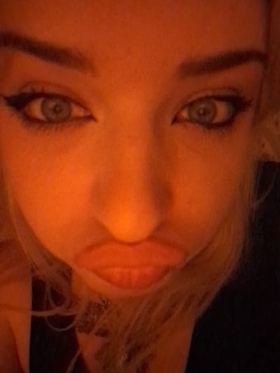 This image captures a close-up, vertically oriented view of a human face, located in Area D (Elaho/Garibaldi), Canada. The subject, an individual with light-colored eyes, features prominent dark winged eyeliner and defined dark, arched eyebrows. The entire face is uniformly illuminated by a dominant orange-red light source. The lips are full, reddish, and intentionally formed into a puckered or 'pout' expression. Strands of light-colored hair are visible framing the lower face. A dark, slender object, potentially a necklace or chain, is faintly visible below the chin against an indistinguishable dark background. No other individuals, objects, or environmental details are present within the frame.