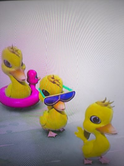 Three yellow cartoon ducklings are shown. One wears sunglasses, another has a pink floatation device around its body, and the third is standing with its wing raised. The ducks have pink feet and orange beaks. The backdrop is a light grey with faint vertical lines. The image is possibly from a television or monitor.