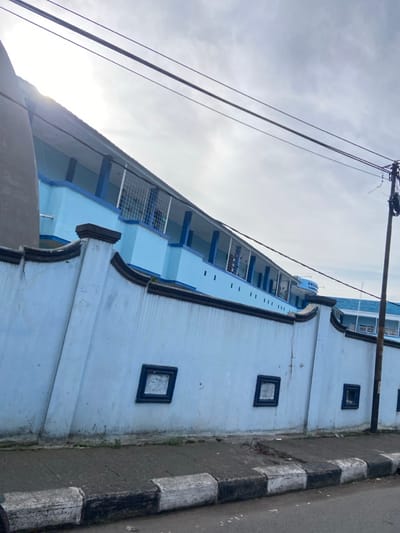 The image shows a blue building with a white fence, a sidewalk, and a street in Margahayu, Indonesia. The building is made of concrete and has a flat roof. The fence is made of concrete and is painted white. The sidewalk is made of concrete and has a black and white checkerboard pattern. The street is paved with asphalt. The sun is shining brightly. The building has multiple rows of windows with black frames and blue trim, and the roof has a blue trim as well. There are two power lines above the building. One of the power lines has an attached utility pole that reaches into the frame of the image. There are no people in the image. The building appears to be abandoned.