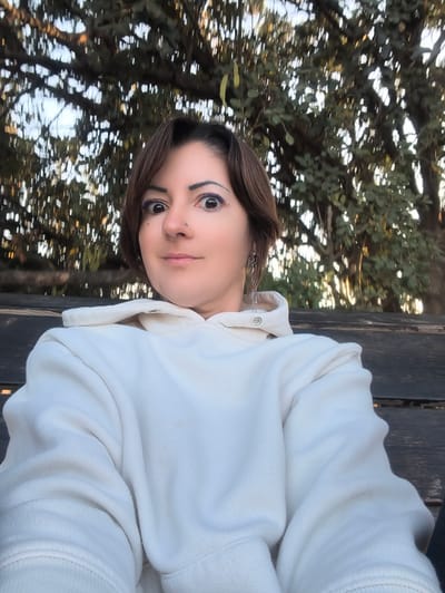 A woman is captured in a close-up, head-on selfie taken in Shaalvim, Israel. She has short, dark brown hair, wide-set eyes with prominent purple eyeliner on both upper and lower lids, and a stud piercing in her right nostril. Her expression features a slight, closed-mouth smile. She is wearing a cream-colored hooded sweatshirt (hoodie) and a silver-toned dangle earring on her left ear. She is seated on a dark, weathered wooden bench, parts of which are visible in the lower half of the frame. The background is an out-of-focus expanse of dense green tree foliage under natural, diffused lighting, indicating an outdoor setting.