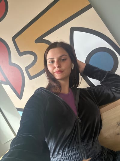 A young woman with dark hair and light skin is taking a selfie in front of a mural. She is wearing a black velour tracksuit and a purple top. She has on white wireless earbuds and is touching her hair with her right hand. The mural behind her is abstract and features large, stylized eye shapes in various colors, including red, yellow, and blue, outlined in black. The setting appears to be indoors, possibly a cafe or a public space, with a wooden surface visible in the foreground. The overall lighting is bright. The location is not specified in the image.