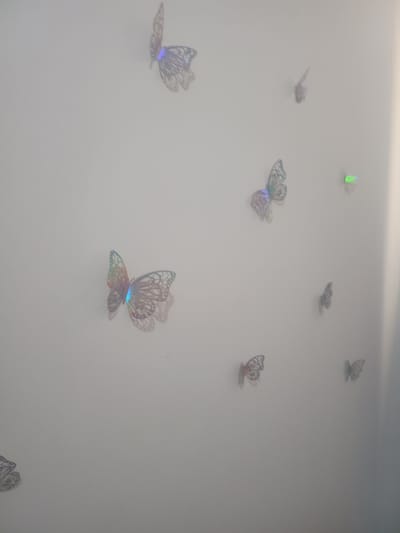 The image displays a wall decorated with numerous iridescent butterfly stickers, predominantly in a light grey or white hue, with some exhibiting a holographic sheen that reflects blue and green light. There are no people present in the scene. The butterflies are adhered to a plain, light-colored wall, suggesting an indoor setting. The composition is from a medium distance, capturing several butterflies of varying sizes scattered across the wall. The lighting appears soft and diffuse, contributing to the slightly hazy quality of the image. This image is not associated with the city of Koryta-Osada, Poland.