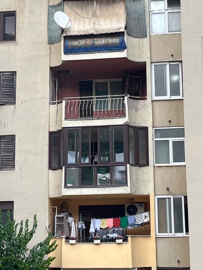 The image shows a close-up, eye-level, vertical shot of an apartment building facade in Škaljari, Montenegro. The building is constructed from light-colored concrete, with visible signs of wear and tear, including some staining and discoloration on the walls. The facade is segmented by balconies and windows, with the central section featuring a prominent bay window with dark brown framing, containing multiple panes. Inside this bay window, laundry is hanging on a line, including several items of clothing and a white sheet. Below this, on a lower balcony, more laundry is hung out to dry, featuring a variety of colored t-shirts, striped garments, and white items, alongside a clothes drying rack. An air conditioning unit is mounted on the wall above this lower balcony.
Several windows with dark brown shutters are visible on the left side of the building. On the right side, there are several windows with white frames. One of these, on the uppermost visible floor, has a satellite dish mounted on the exterior. The upper part of the building displays a section of weathered, off-white fabric awning supported by a corrugated metal structure. At the very top of the image, a white satellite dish is prominently positioned. Green foliage from a tree or bush encroaches on the lower left corner of the frame. The scene appears to depict a residential area, possibly in an urban or suburban setting, with everyday life activities like laundry drying being observed.