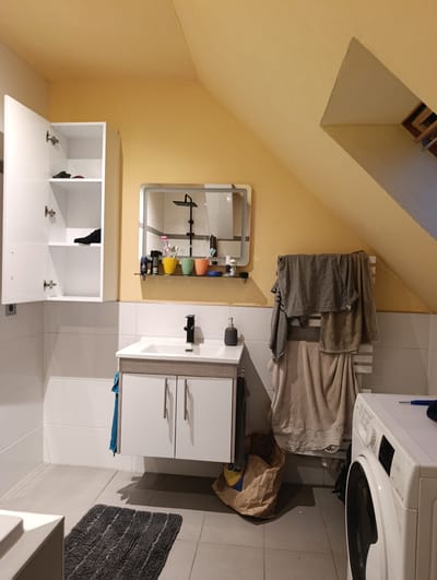 The image shows a bathroom in Roßlau, Germany. The bathroom is relatively small and has an angled ceiling painted yellow. The walls are tiled in white. There is a white vanity with two drawers and a grey countertop, with a white sink and a black faucet. Above the sink is a large mirror with an integrated light. On the mirror shelf are several toiletries, including toothbrushes in colored cups, deodorant, and bottles.
To the left of the vanity, there is a tall, narrow white cabinet with its door open, revealing two shelves with a black shoe and a red item on them. A blue towel is hanging from the side of the vanity.
To the right of the vanity, a white towel rack is mounted on the wall, with several folded towels and a grey t-shirt hanging on it. A brown paper bag is on the floor next to the towel rack. In the foreground on the right, a white washing machine is visible, with its door slightly ajar. A dark blue object, possibly a cleaning cloth or sponge, rests on top of the washing machine. A dark grey bath mat is on the floor in front of the vanity. The floor is tiled with large grey tiles. The overall impression is a functional but somewhat cluttered bathroom.