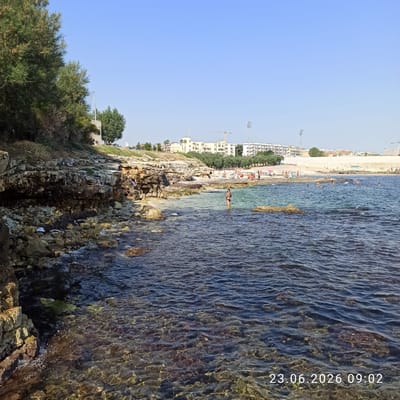 A coastal scene in Molfetta, Italy, features a rocky shoreline with a clear blue sky overhead. The water is a vibrant turquoise near the shore, gradually deepening to a darker blue. In the water, several people are visible, some wading and others swimming. A woman in a black bikini stands in the shallows. Further out, a group of people are sunbathing on a sandy beach, backed by trees and buildings that suggest an urban or resort area. The rocky coastline is covered in patches of vegetation and seaweed. The image is dated "23.06.2026 09:02."
